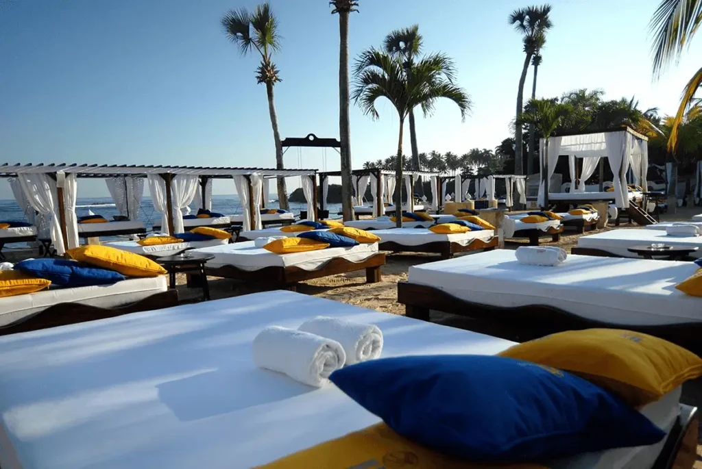 "A pristine beach at a luxury resort features rows of white cabanas and daybeds with blue and yellow pillows, and tall palm trees under a clear blue sky, inviting guests to relax and explore amazing fox travel deals".