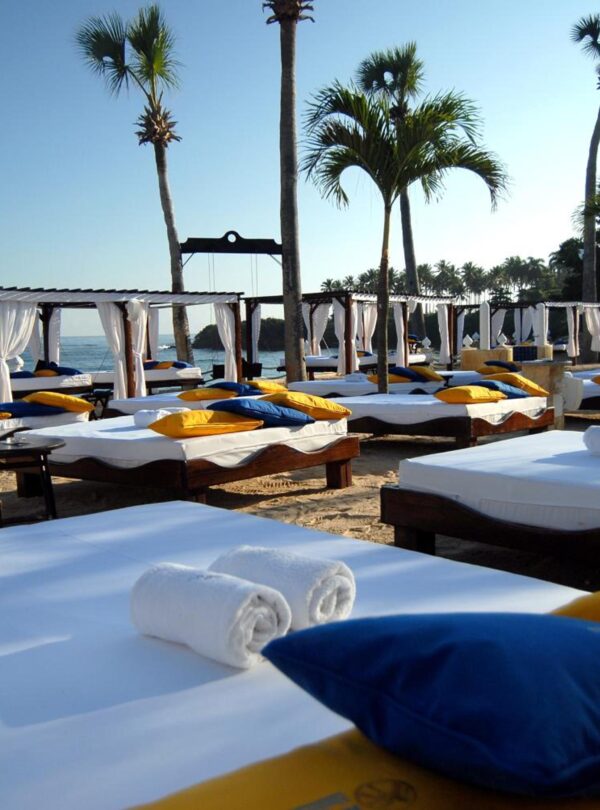 Row of luxurious white beach cabanas with blue and yellow pillows on a sandy beach under palm trees at Lifestyle Tropical Beach Resort & Spa, with the ocean and distant land visible under a clear blue sky.